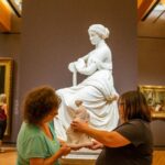 two woman use a touchable miniature of a statue of a seated woman in a museum