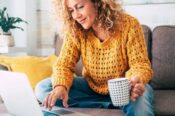 young caucasian woman looking at laptop with coffee mug in one hand while sitting on sofa
