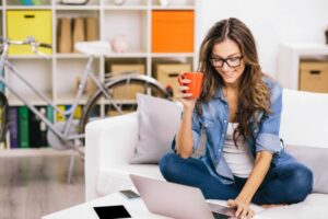 young caucasian woman looking at laptop with coffee mug in one hand while sitting on sofa2